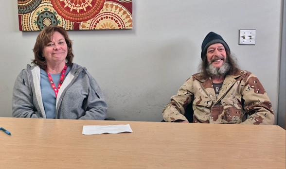 A woman in a gray sweatshirt sits next to a man in a winter hat and camo coat at a table