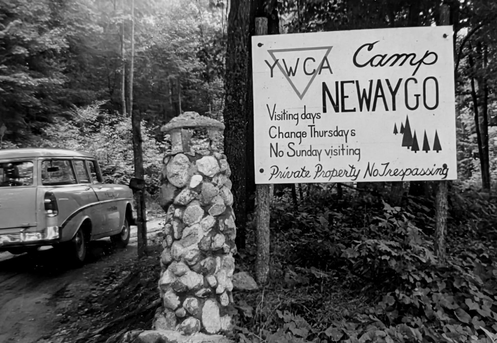 An old black and white photo of a 1950's-era station wagon driving past the old YWCA Camp Newaygo sign and stone gate.